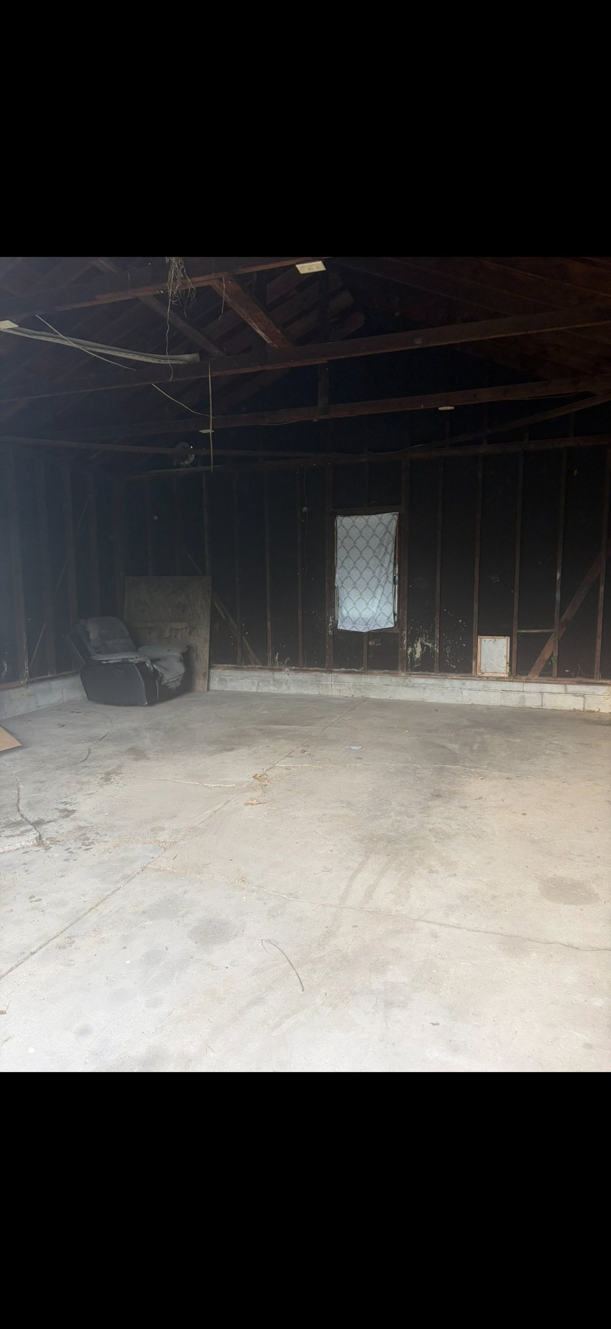 Interior of a dimly lit garage with concrete floor, dark wooden structure, metal door with wire mesh window, and power lines visible overhead